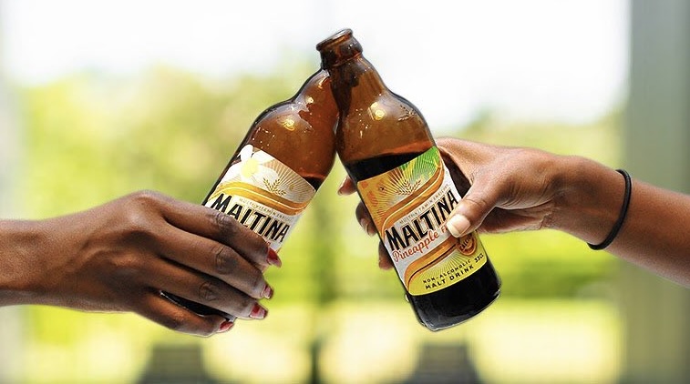 Two hands clinking two bottles of Maltina, a non-alcoholic soft drink.