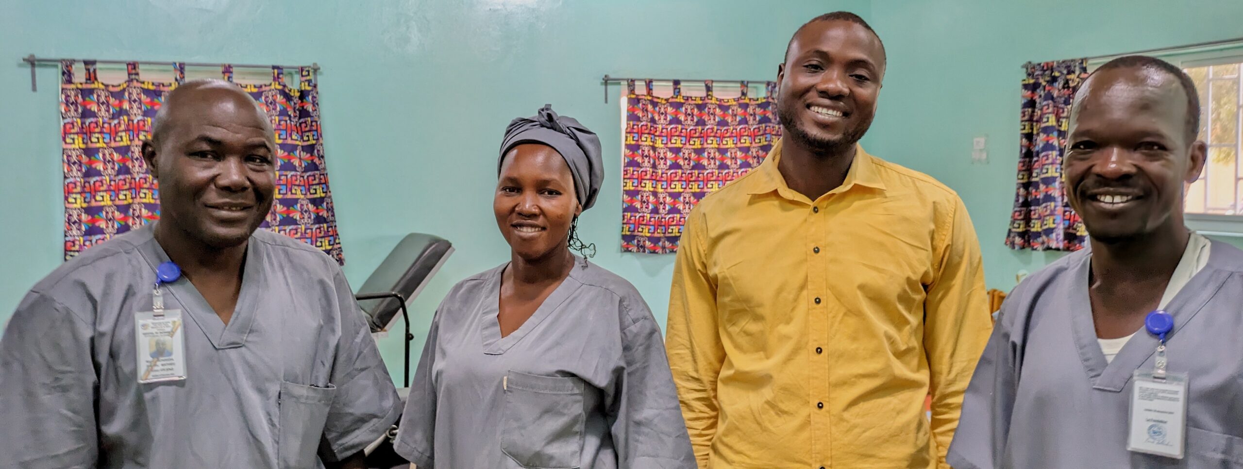 Three men and a woman stand in a row. They all wear grey medical scrubs apart from one man in a yellow shirt. This is the team helping to care for children at the child development centre. From left to right: Mathieu, Cria, Osée, Jean.