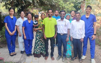 A group of people stand in a garden in India. Many of them are medics wearing blue scrubs. In the centre is a young man, Lakhan, wearing a green shirt. He stands next to Dr Jibi John who wears a white shirt. He also stands next to his wife who is wearing a sari, and holding a small boy, one of their children.