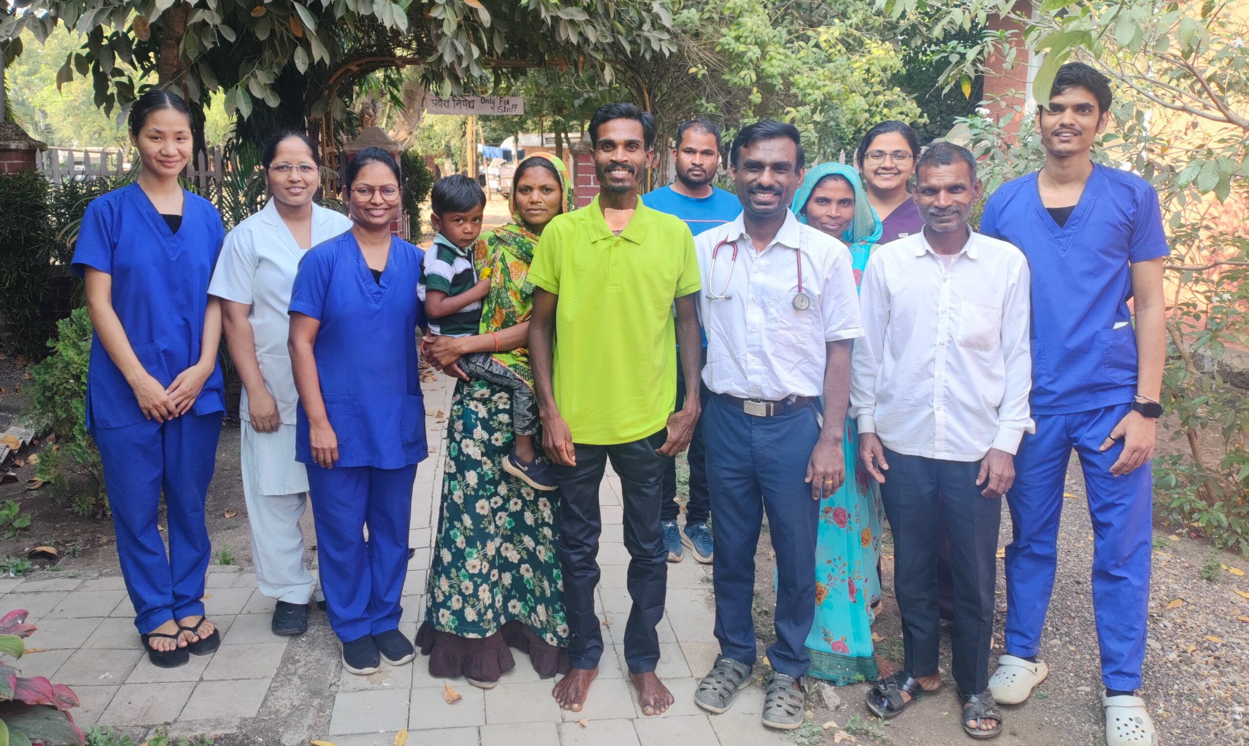 A group of people stand in a garden in India. Many of them are medics wearing blue scrubs. In the centre is a young man, Lakhan, wearing a green shirt. He stands next to Dr Jibi John who wears a white shirt. He also stands next to his wife who is wearing a sari, and holding a small boy, one of their children.