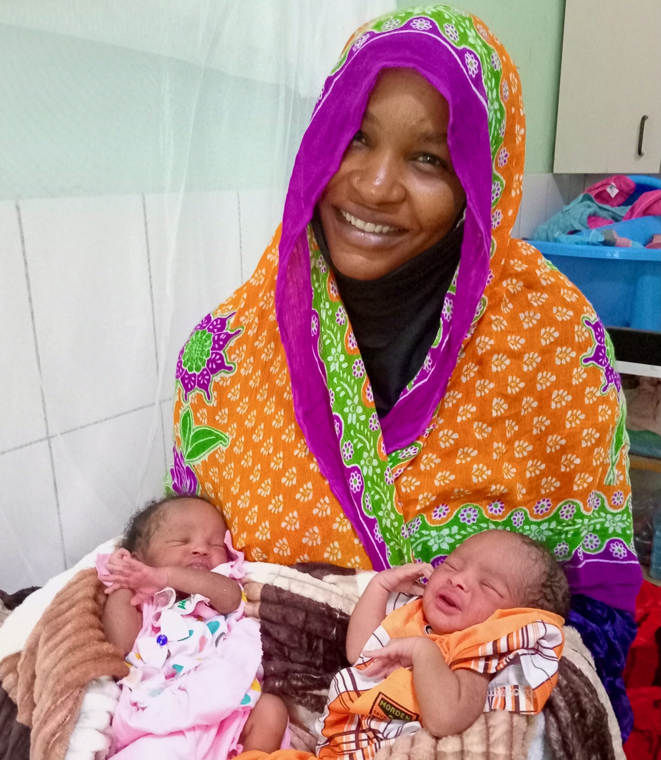 A lady from Chad smiles holding her twins in her lap.