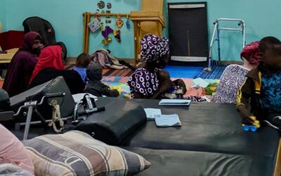 A group session at the child development centre at the Guinebor 2 Hospital in Chad. A group of women and children sit in a blue hospital room with toys and therapy equipment including beds and walkers.